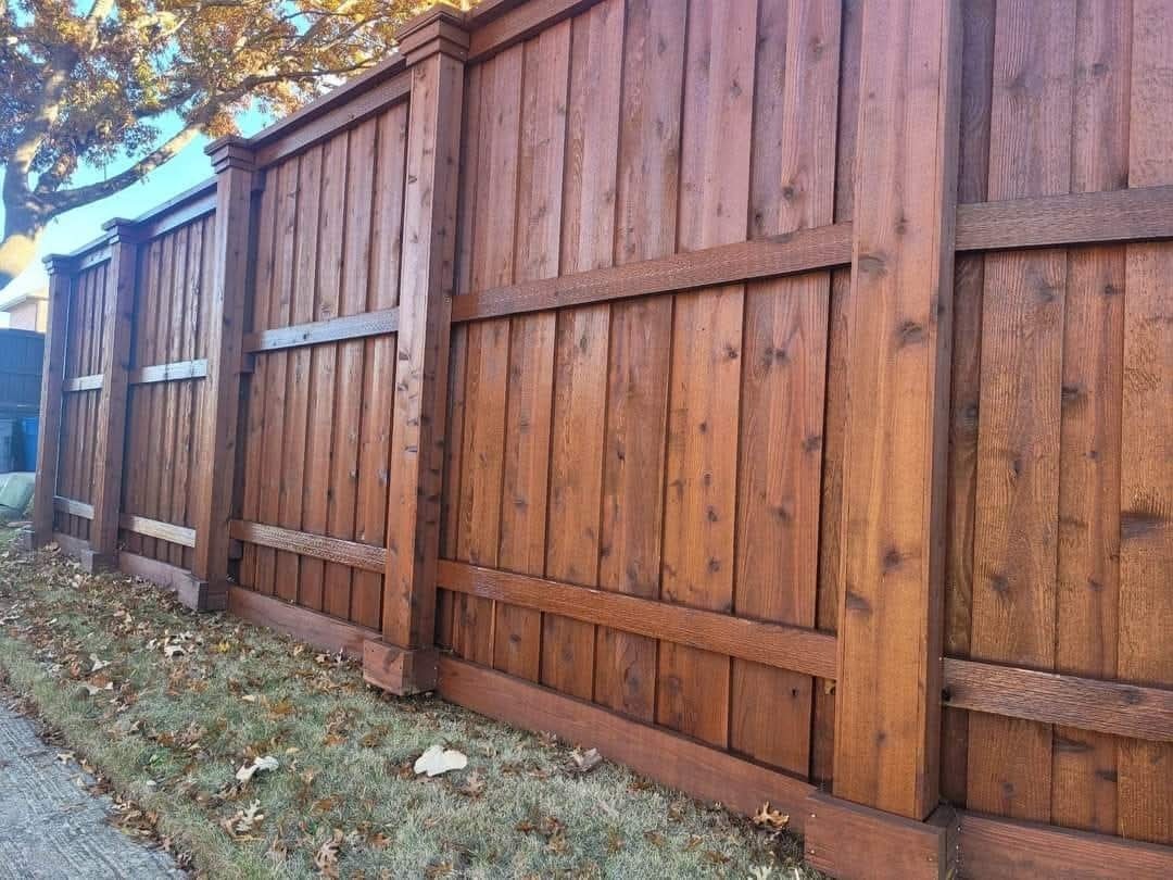 A stained wood fence turning a corner under a shade tree with fall leaves on the ground