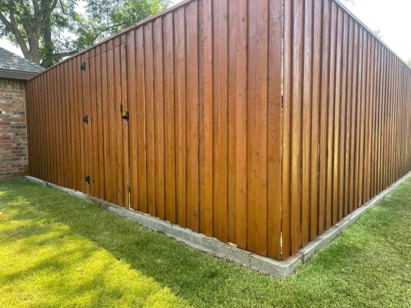 A reddish brown stained wood fence and gate beside a brick home with a lush lawn