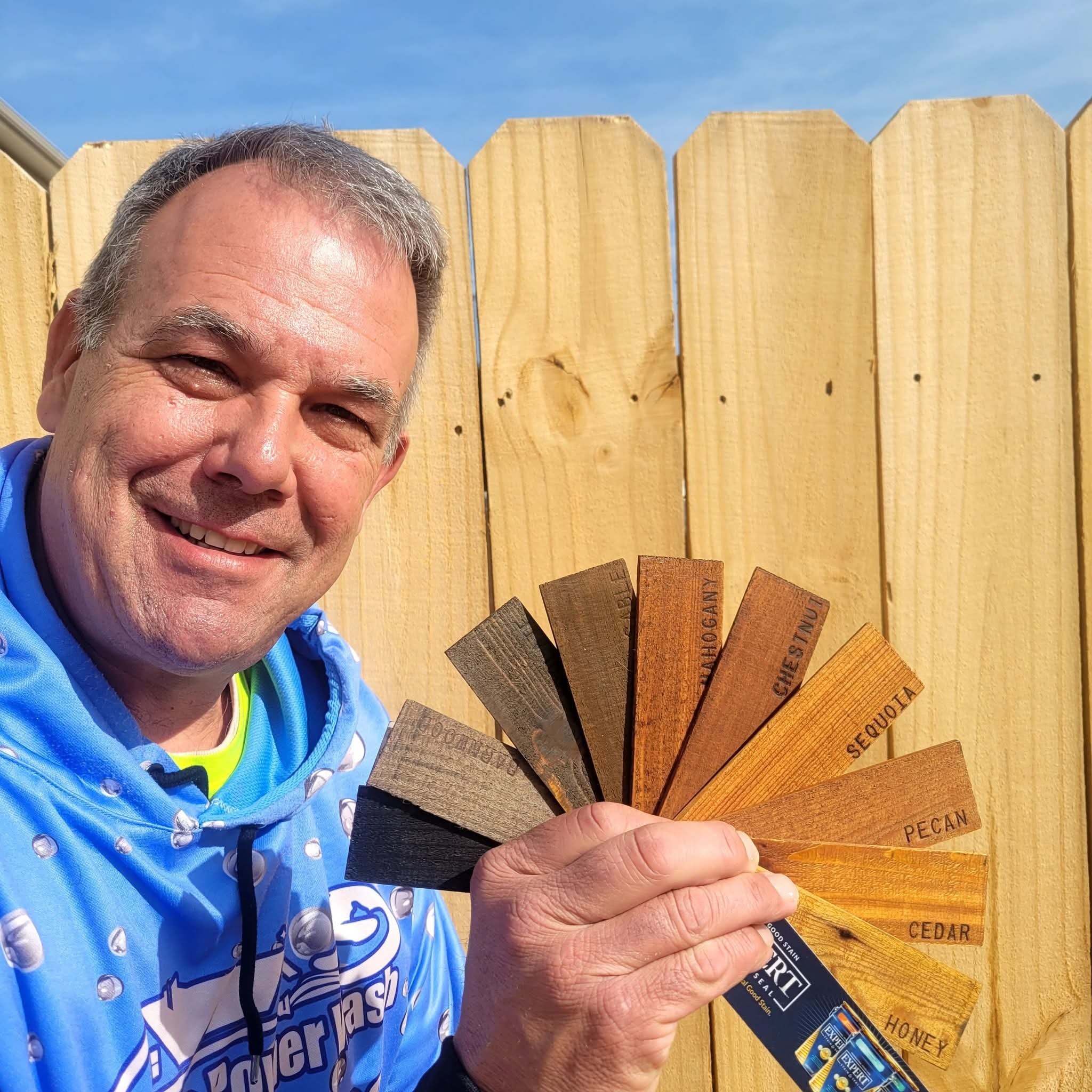 A crew member holding a wood stain color sample fan in front of a natural wood fence