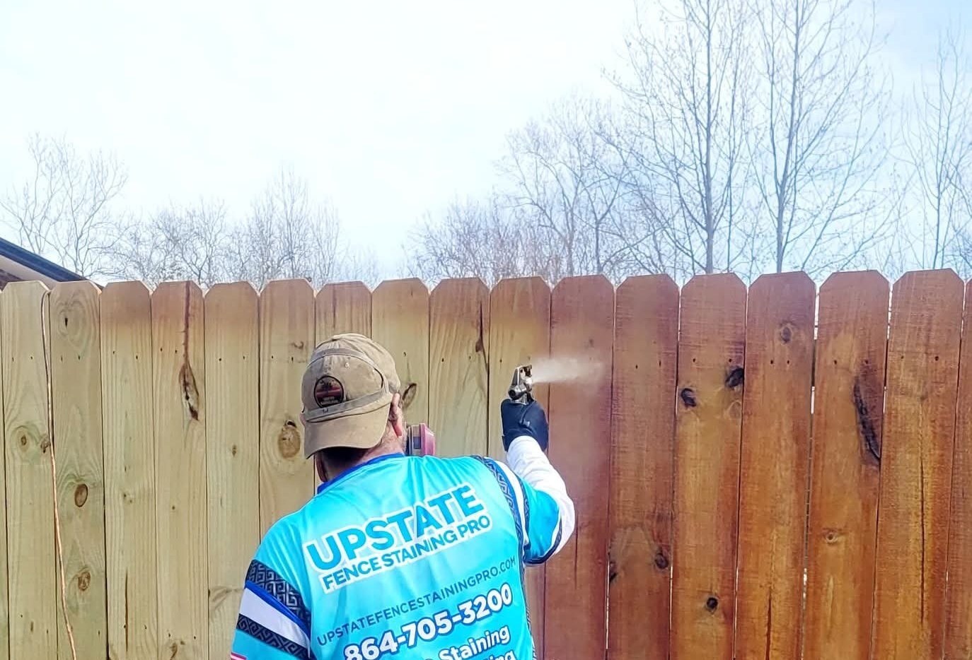 A crew member in an Upstate Fence Staining jersey spraying stain onto a wood privacy fence, natural wood on one side and freshly stained wood on the other